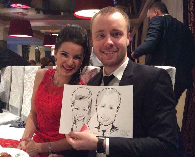 шаржит на свадьбе в Минске Девушка в красном и мужчина улыбаясь держат шарж на свадьбе в Променаде Минск